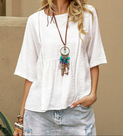 Person wearing a white blouse with a long necklace against a beige wall.
